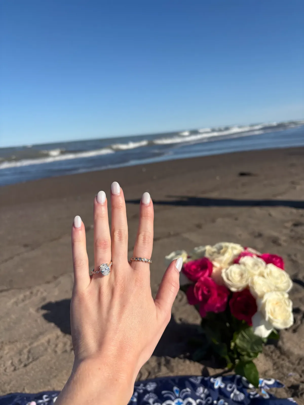 Joshua Nicolls' fiancée showing her oval Joseph-Anthony engagement ring and stacked band on the beach beside a bouquet of roses